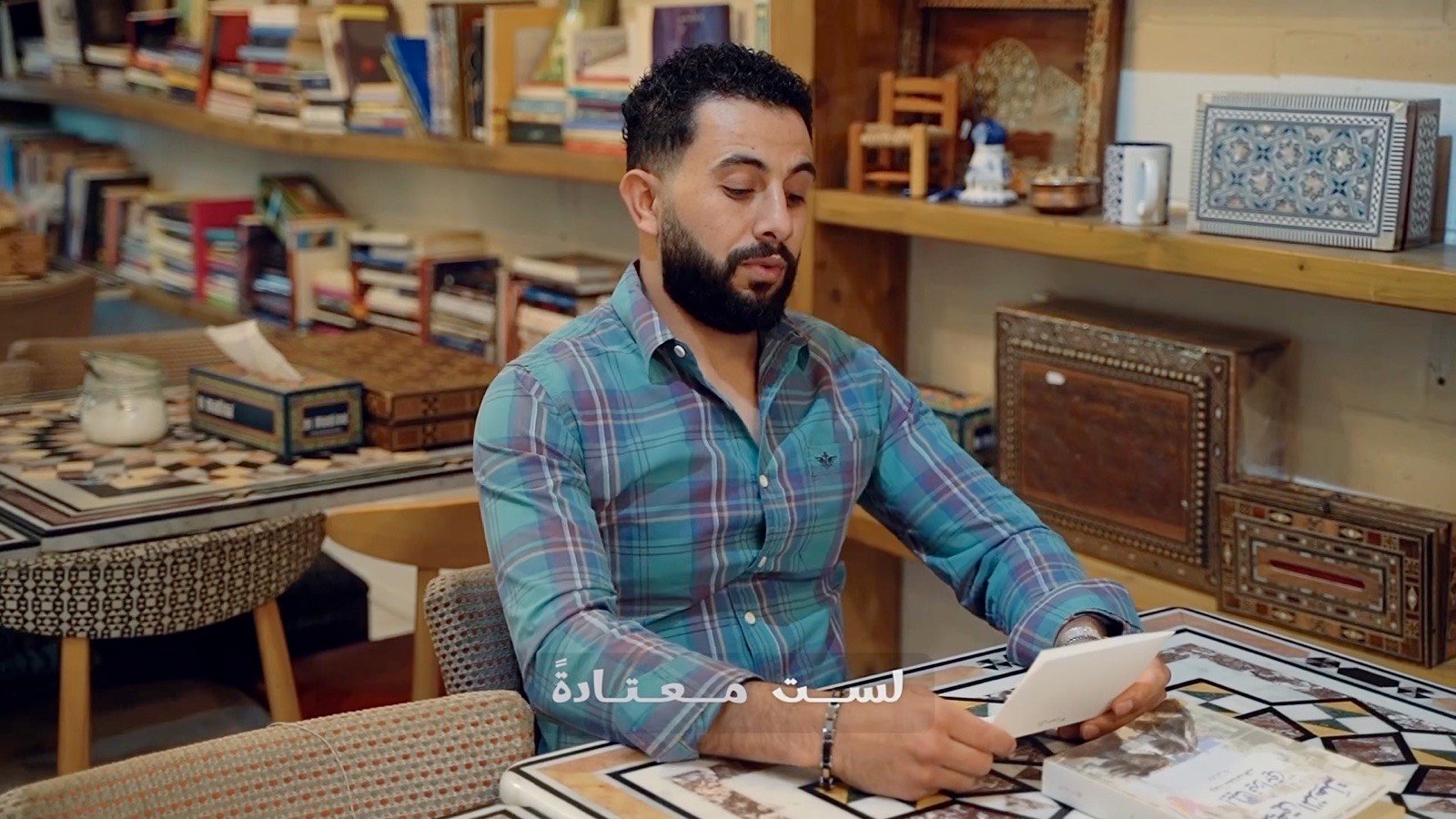 A man in a checked shirt writing at a mosaic table in a café lined with books