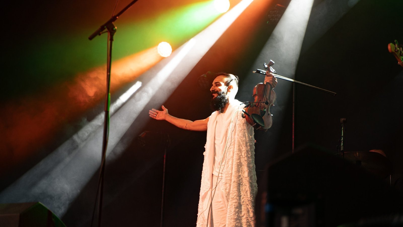 A violinist in white with arms open under stage spotlights