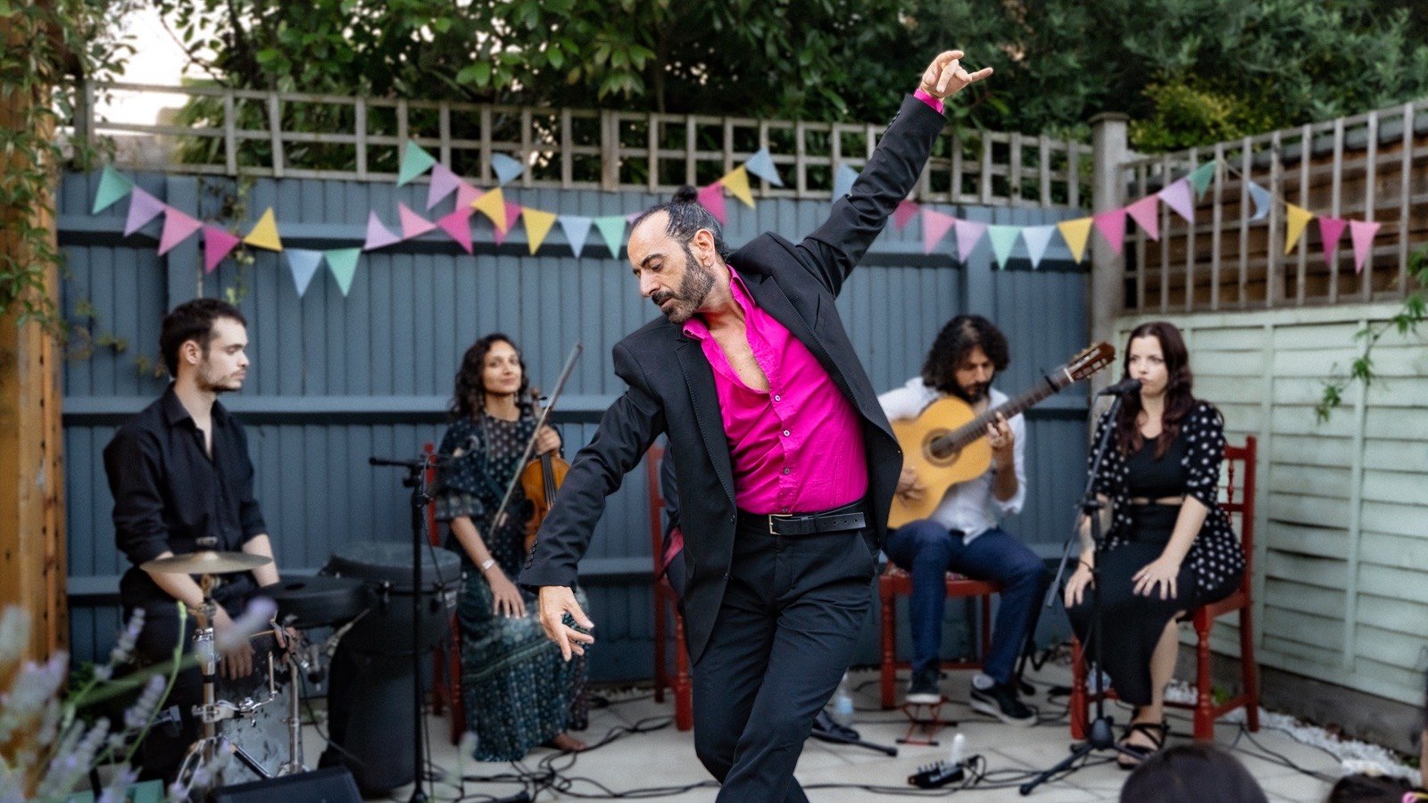 A dancer in a pink shirt mid-turn in front of a band in a garden hung with bunting