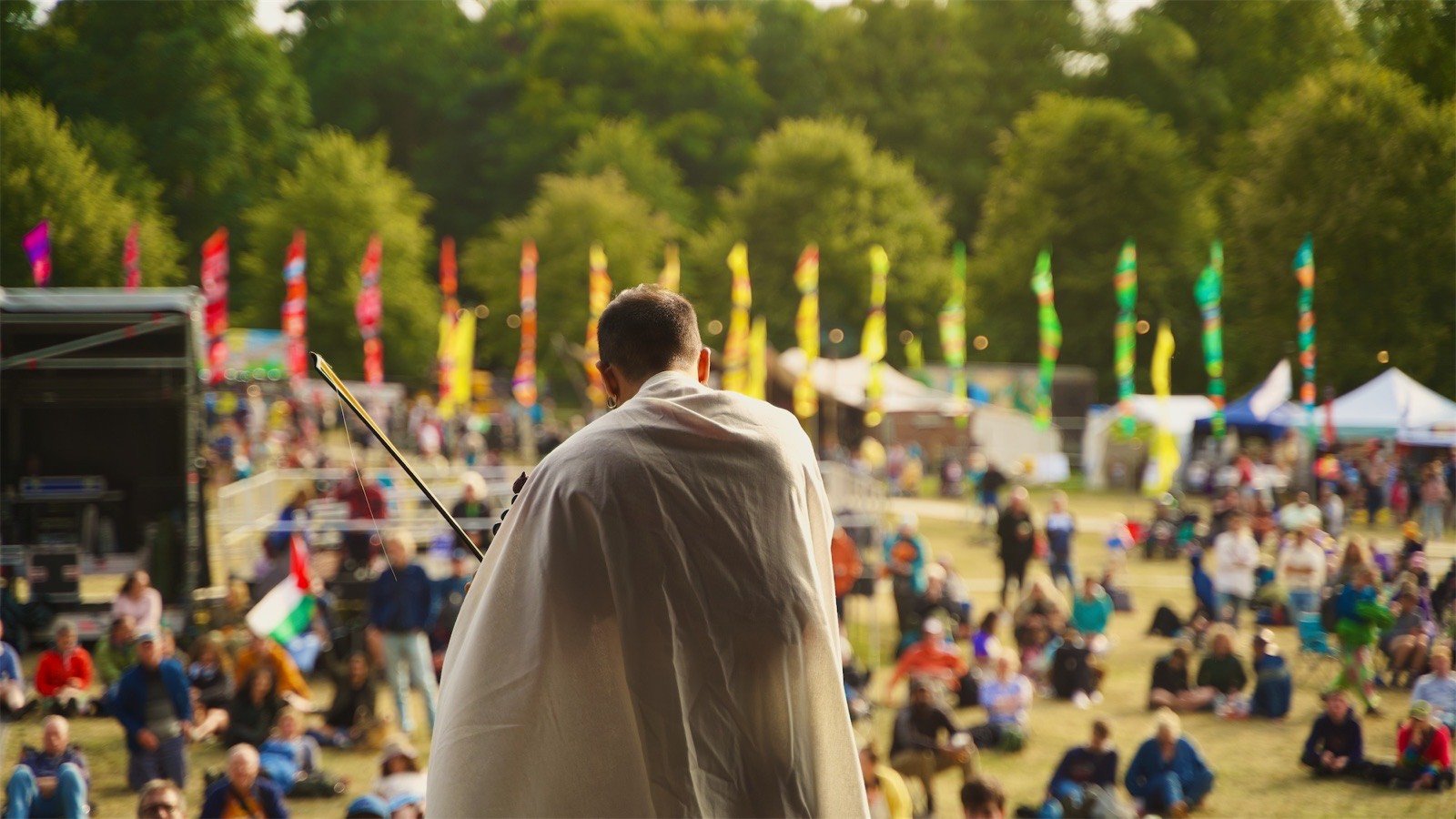 A violinist in white, seen from behind on stage, facing a festival crowd under rows of flags