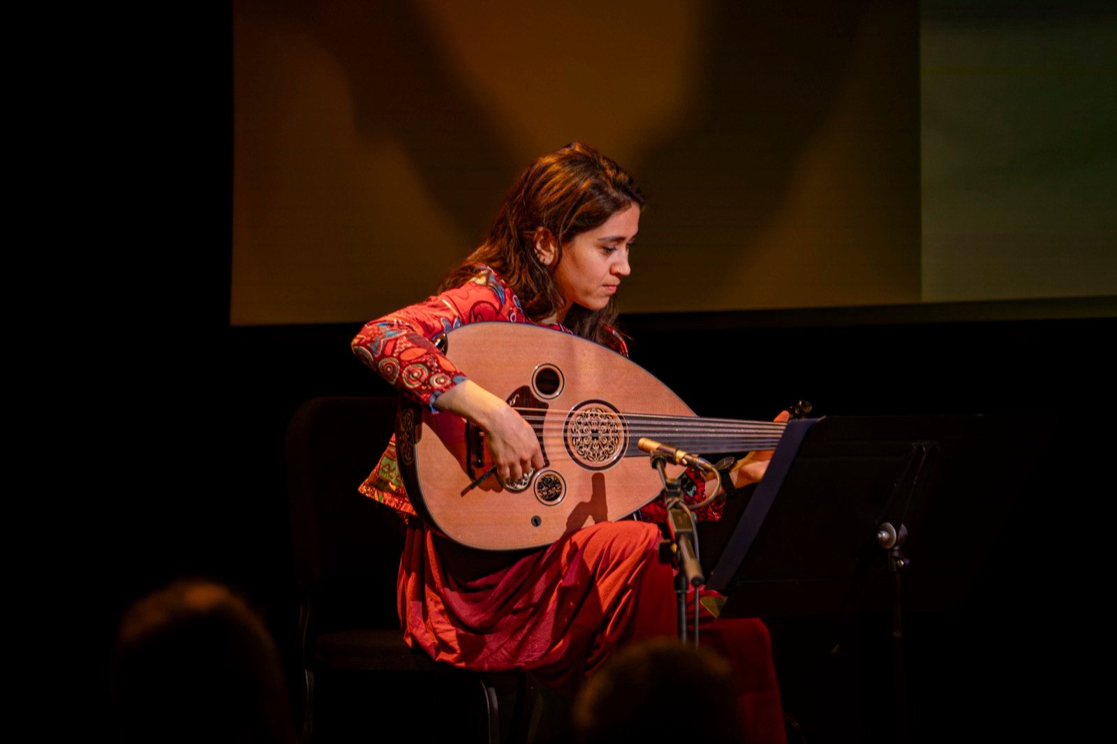 An oud player in a red embroidered dress playing on a dark stage