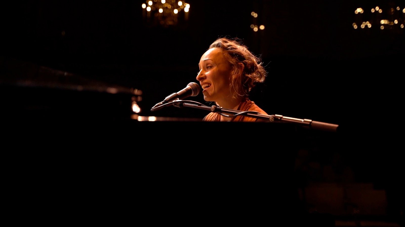 A singer at a grand piano under chandeliers
