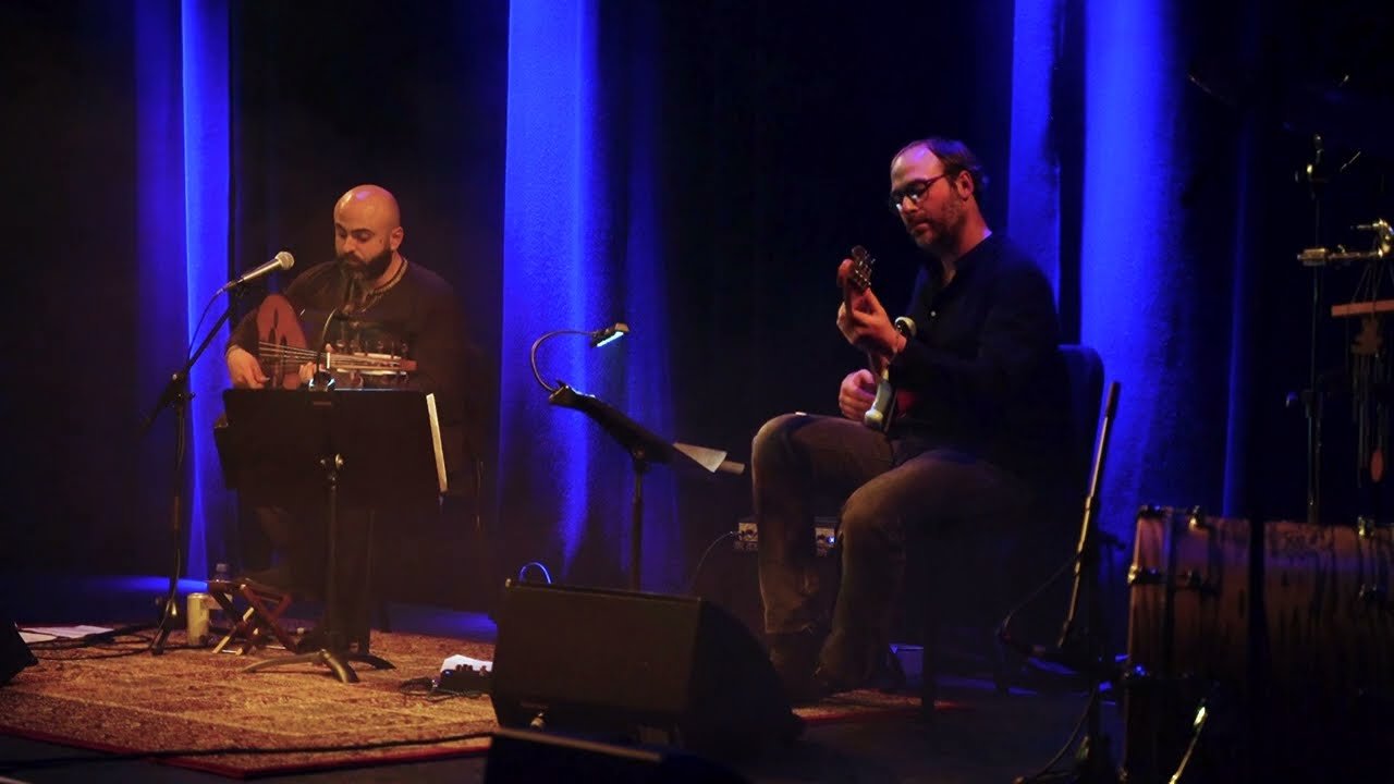 An oud player and a guitarist on a stage lit blue