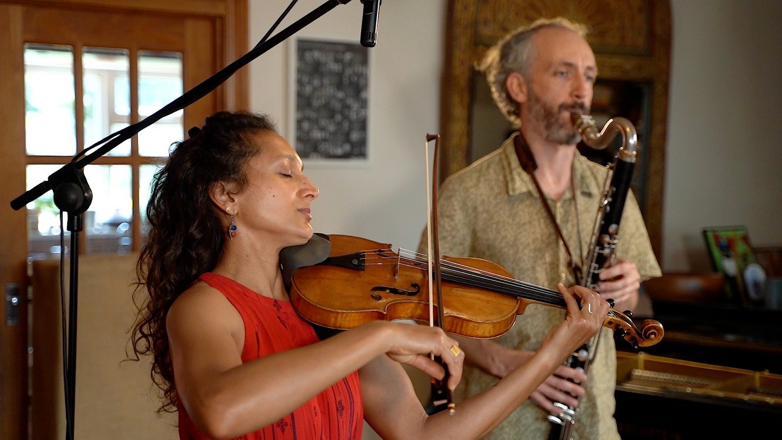 A violinist in red and a bass clarinettist playing together in a wood-panelled room